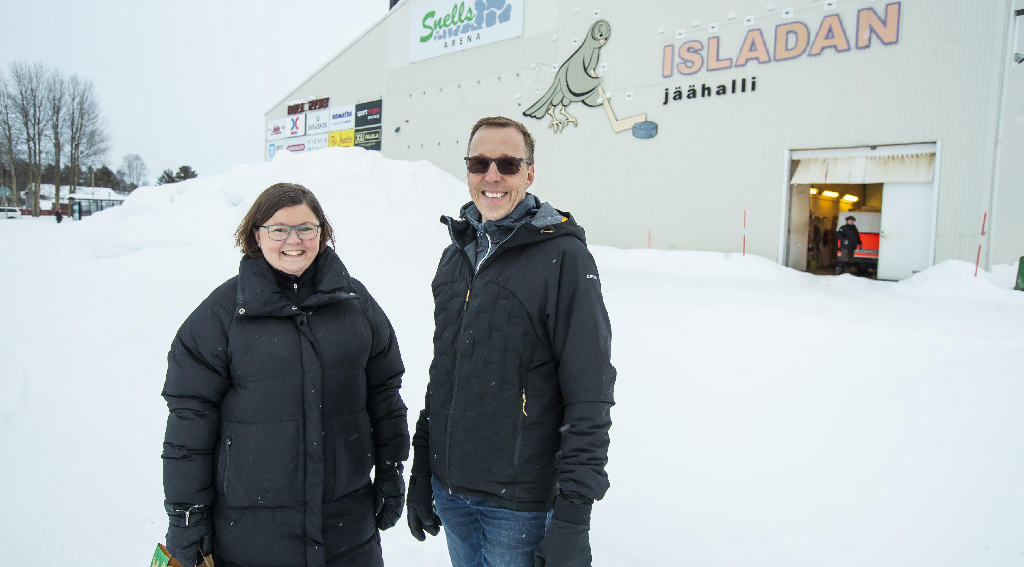 En kvinna och en man står i snön utomhus framför en ishall