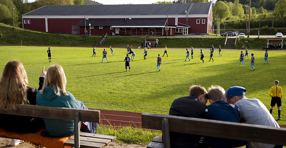 En fotbollsplan med åskådare i förgrunden och byggnader i bakgrunden