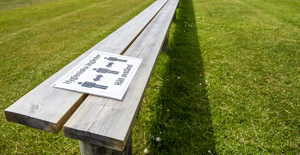 Bild på en bänk med skyltning om att hålla avstånd, vid en fotbollsplan