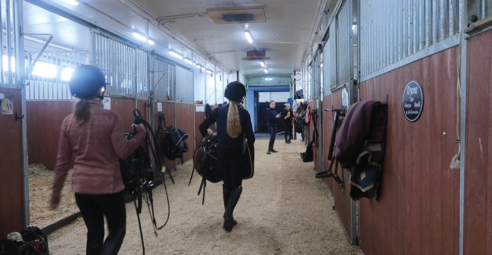 Två unga tjejer med ryggarna mot kameran går och bär på varsin hästsadel i ett stall. 