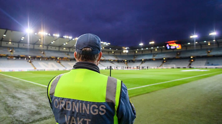 En ordningsvakt blickar ut över en upplyst fotbollsplan inne på en arena. 
