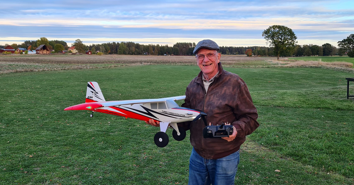 En man står utomhus på flygfältet och ser mot kameran med ett modellflygplan i handen