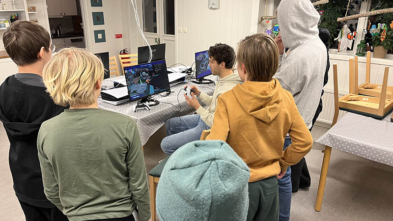 En vuxen sitter vid en datorskärm och visar hur det går till att spela. En grupp unga stån och tittar med ryggen mot kameran.