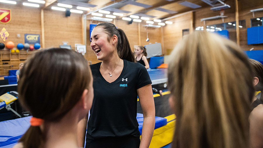 En ung kvinna som är ledare står framför flickor i en gympasal. Hon skrattar åt något. 