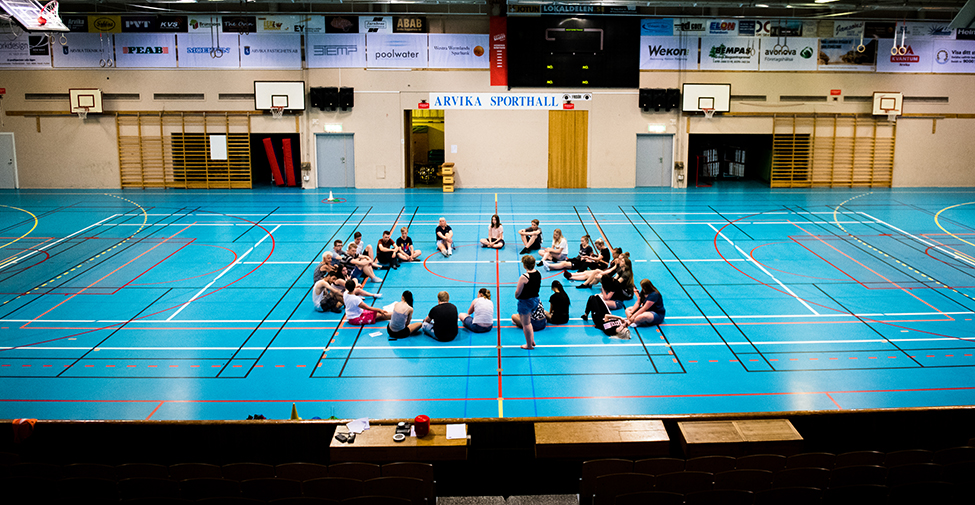 En grupp idrottsaktiva sitter i en cirkel på golvet till en idrottshall