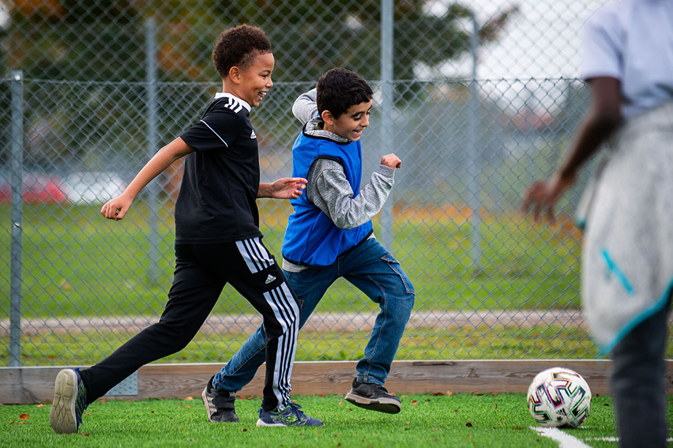 Två barn spelar fotboll på gräsplan utomhus.