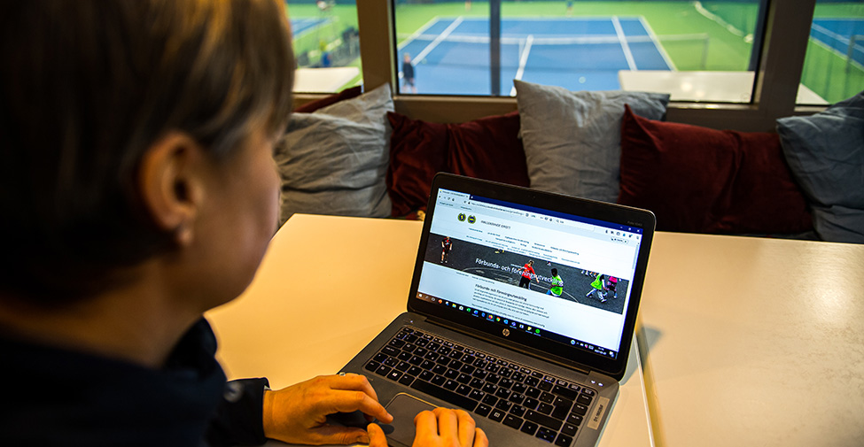 En kvinna läser på skärmen på en bärbar dator. Hon sitter inne i en idrottshall. I bakgrunden syns en tennisplan. 